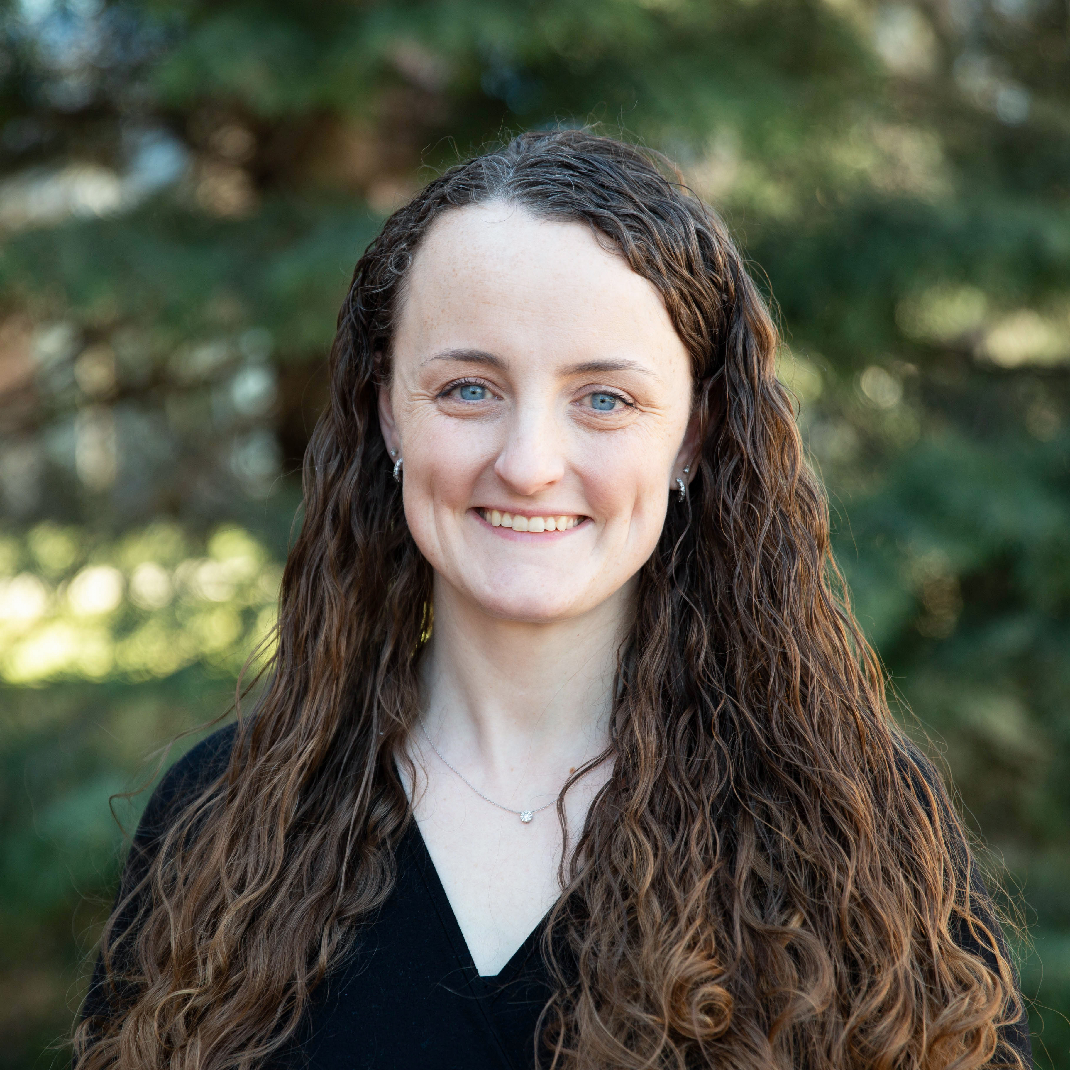 A photo of a woman with long wavy brown hair, wearing a black v-neck blouse against a tree lined background. 