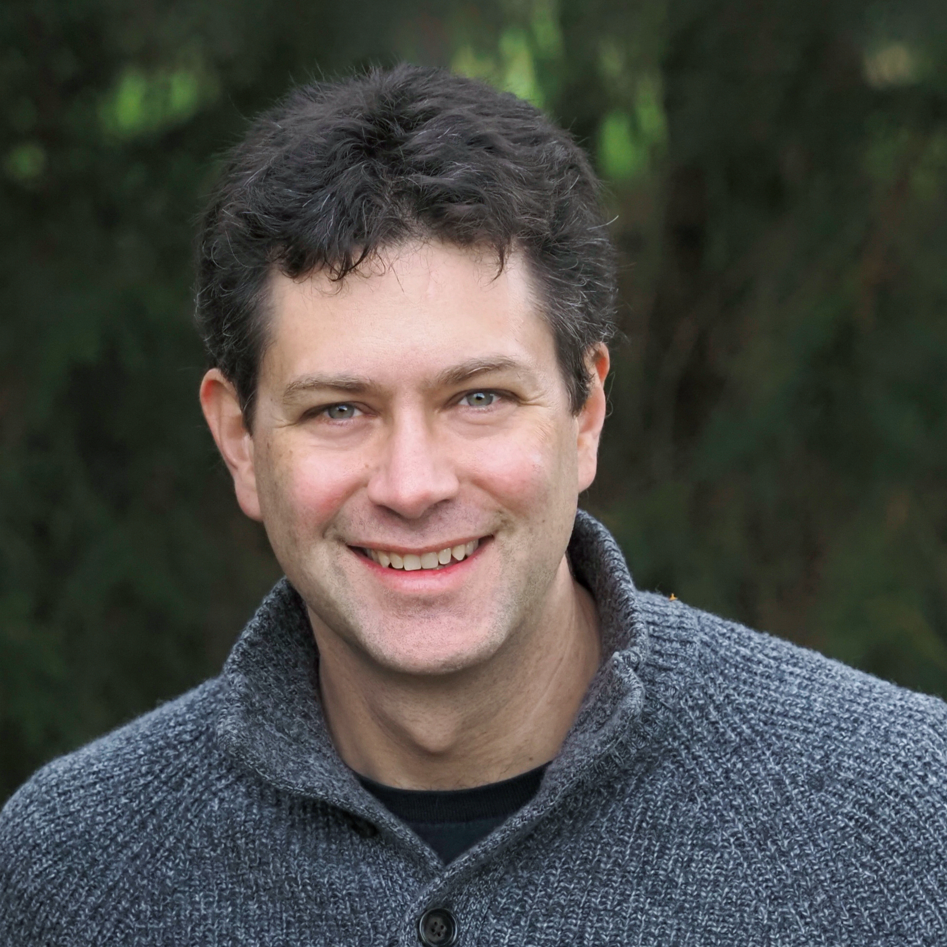 A man with short dark hair in a grey sweater, against a tree lined background