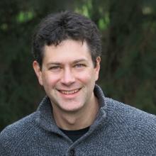 A man with short dark hair in a grey sweater, against a tree lined background