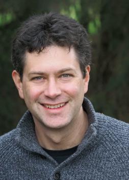 A man with short dark hair in a grey sweater, against a tree lined background